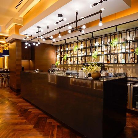 ホテル ジャバール オマール ハイアット リージェンシー マカー メッカ エクステリア 写真 The photo shows a modern bar area in a restaurant. The bar features a sleek, dark countertop and is backed by a decorative arrangement of shelves that holds various bottles and plants. The lighting is warm and stylish, with multiple hanging pendant l