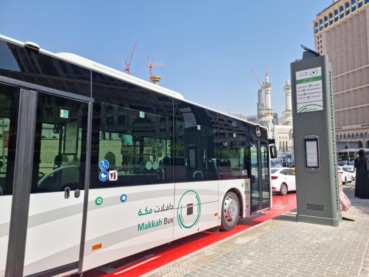 ホテル ジャバール オマール ハイアット リージェンシー マカー メッカ エクステリア 写真 The photo shows a public bus labeled "Makkah Bus," parked at a designated bus stop. The bus has a modern design and is predominantly white, with green accents. In the background, there are tall buildings and construction cranes, indicating a busy urb