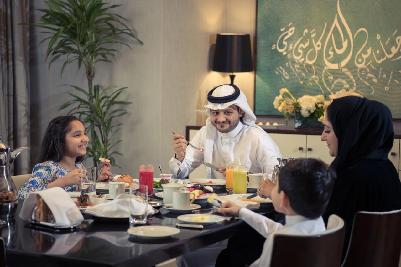 ホテル ジャバール オマール ハイアット リージェンシー マカー メッカ エクステリア 写真 The photo depicts a cheerful family gathered around a dining table for a meal. They appear to be enjoying breakfast or a brunch. The table is set with various dishes and drinks, including juices and plates of food. 

A man wearing traditional attire 