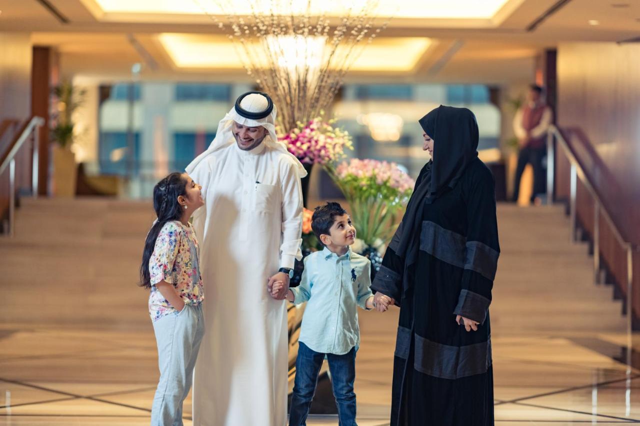 ホテル ジャバール オマール ハイアット リージェンシー マカー メッカ エクステリア 写真 The photo shows a family in a hotel lobby. There are two children, a boy and a girl, standing between their parents. The girl is smiling and looking up at her father, who is wearing traditional white attire and a headscarf. The boy is also smiling an