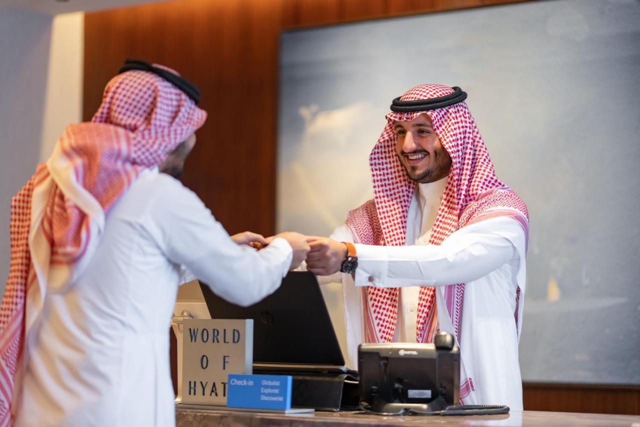 ホテル ジャバール オマール ハイアット リージェンシー マカー メッカ エクステリア 写真 The photo shows a scene at a hotel reception desk. A hotel staff member, dressed in traditional attire, is smiling and handing something to a guest, who is also wearing traditional clothing. The guest appears to be receiving a key or a document. The 