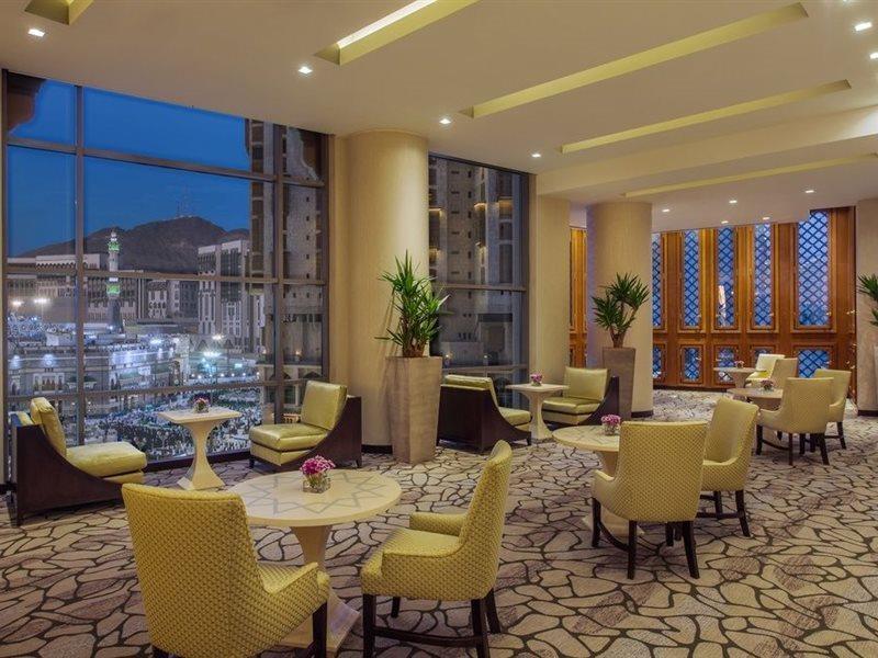 ホテル ジャバール オマール ハイアット リージェンシー マカー メッカ エクステリア 写真 The photo shows a modern, well-lit lounge or reception area. It features several small round tables surrounded by comfortable chairs, all in a soft yellow upholstery. The flooring is composed of stone tiles, adding a natural element to the decor. Lar