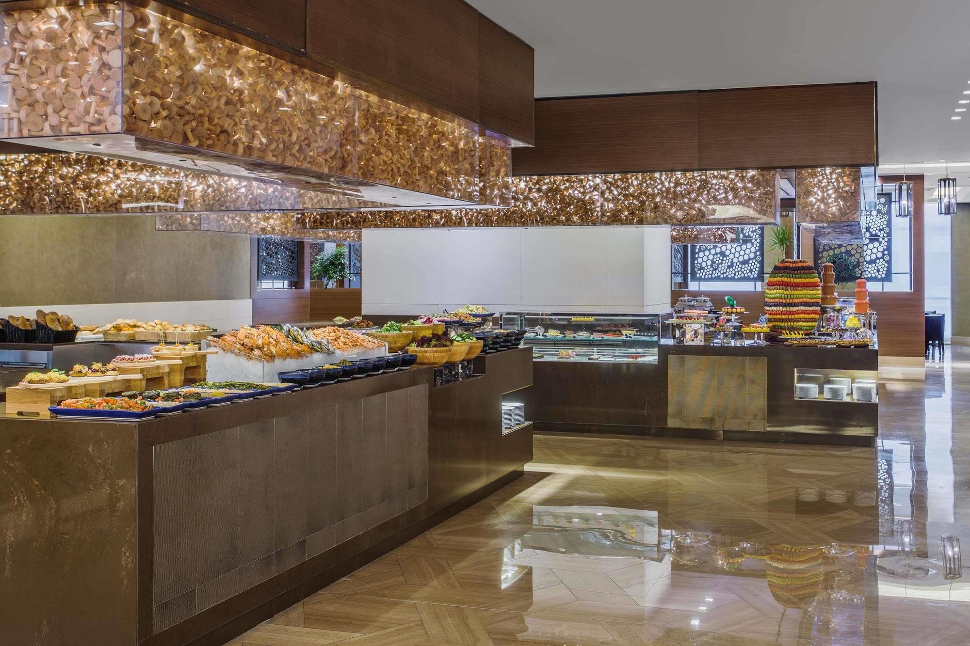ホテル ジャバール オマール ハイアット リージェンシー マカー メッカ エクステリア 写真 The photo shows an elegant buffet setup in a spacious dining area. There are various food displays arranged on sleek surfaces, including an assortment of items such as pastries, fruits, and possibly other dishes. The decor features warm lighting with