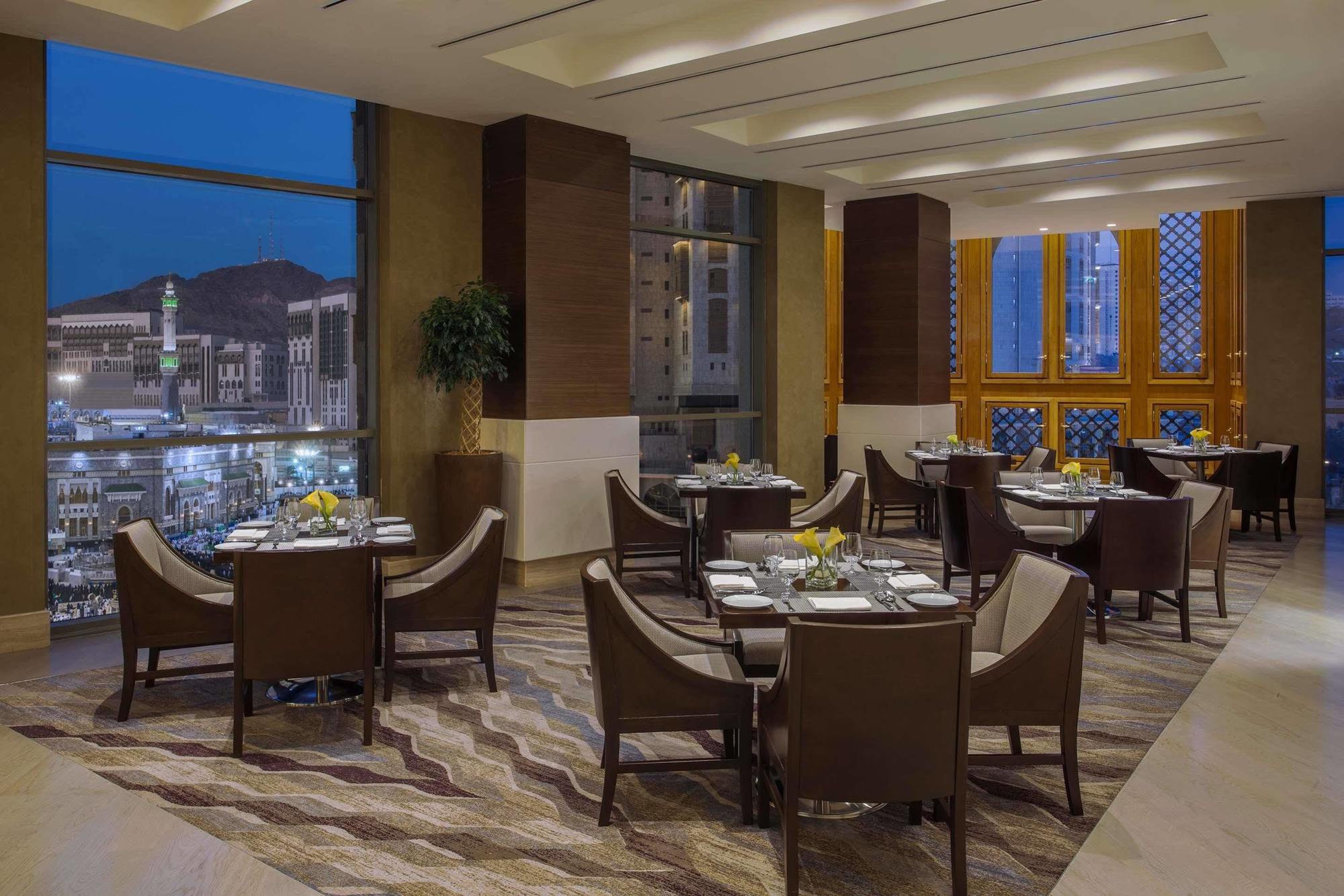ホテル ジャバール オマール ハイアット リージェンシー マカー メッカ エクステリア 写真 The photo shows a sophisticated restaurant interior, featuring elegant round tables with high-backed chairs arranged neatly. The space is well-lit with large windows that provide a view of the city skyline at dusk, with mountains in the background. T