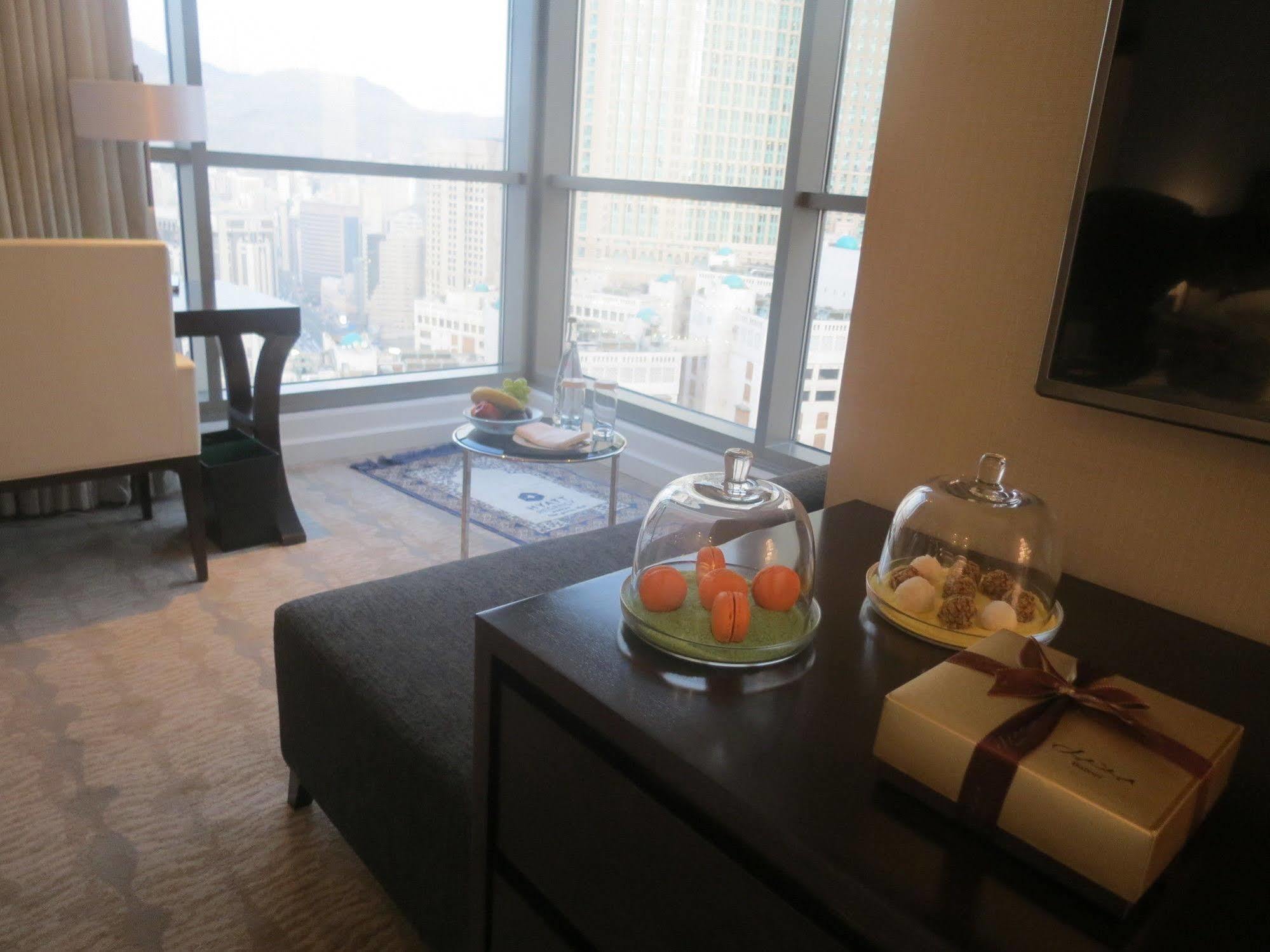 ホテル ジャバール オマール ハイアット リージェンシー マカー メッカ エクステリア 写真 The photo shows a modern hotel room or a stylish living space with large windows offering a view of the city. In the foreground, there is a dark sofa and a low table with covered trays displaying what appears to be small desserts or delicacies. To th