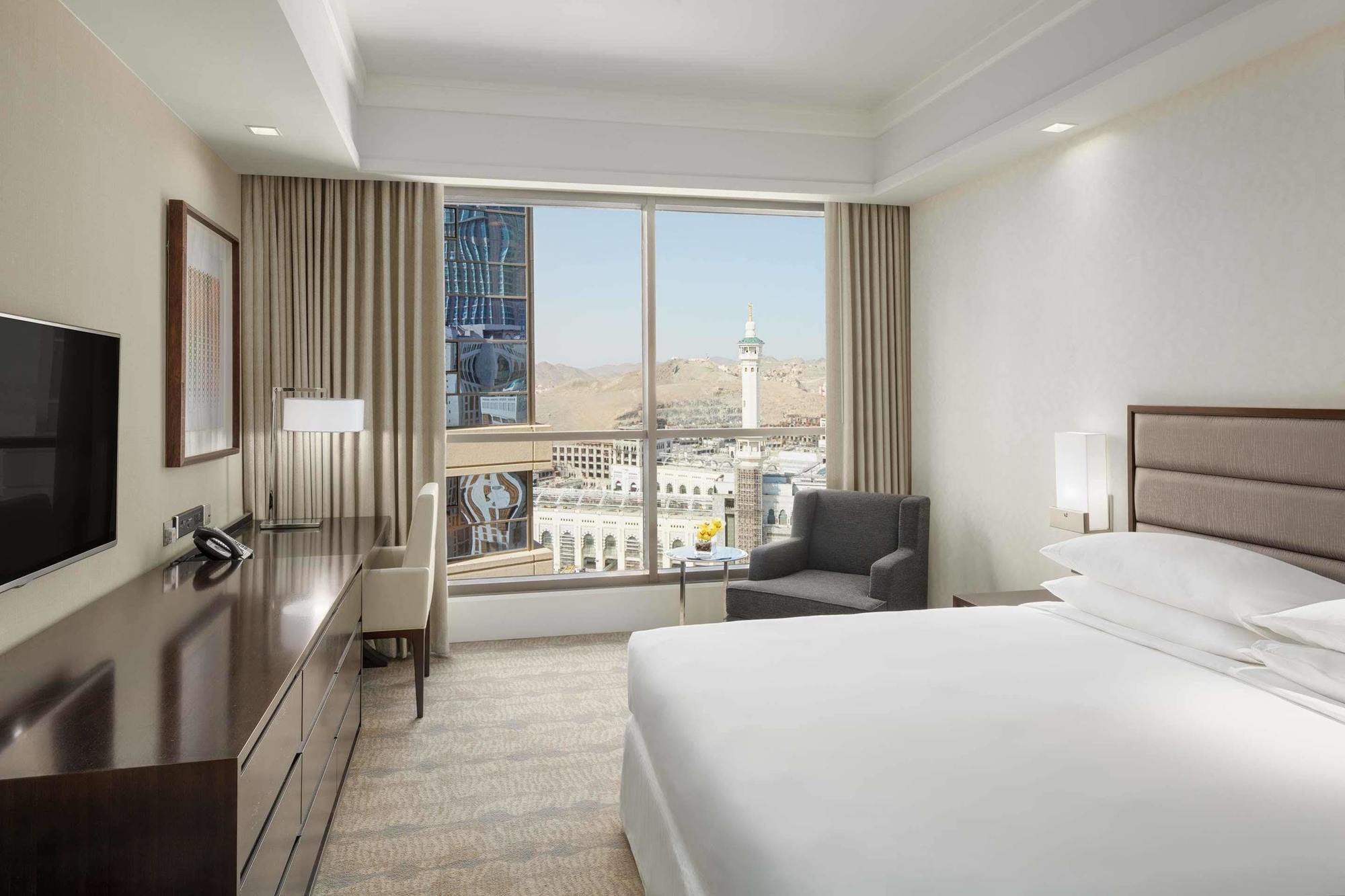 ホテル ジャバール オマール ハイアット リージェンシー マカー メッカ エクステリア 写真 The photo shows a modern hotel room featuring a large window that provides a view of an outdoor landscape, likely part of a city or landmark. The room includes a bed with white linens, a side table with a lamp, and a small seating area with a chair a