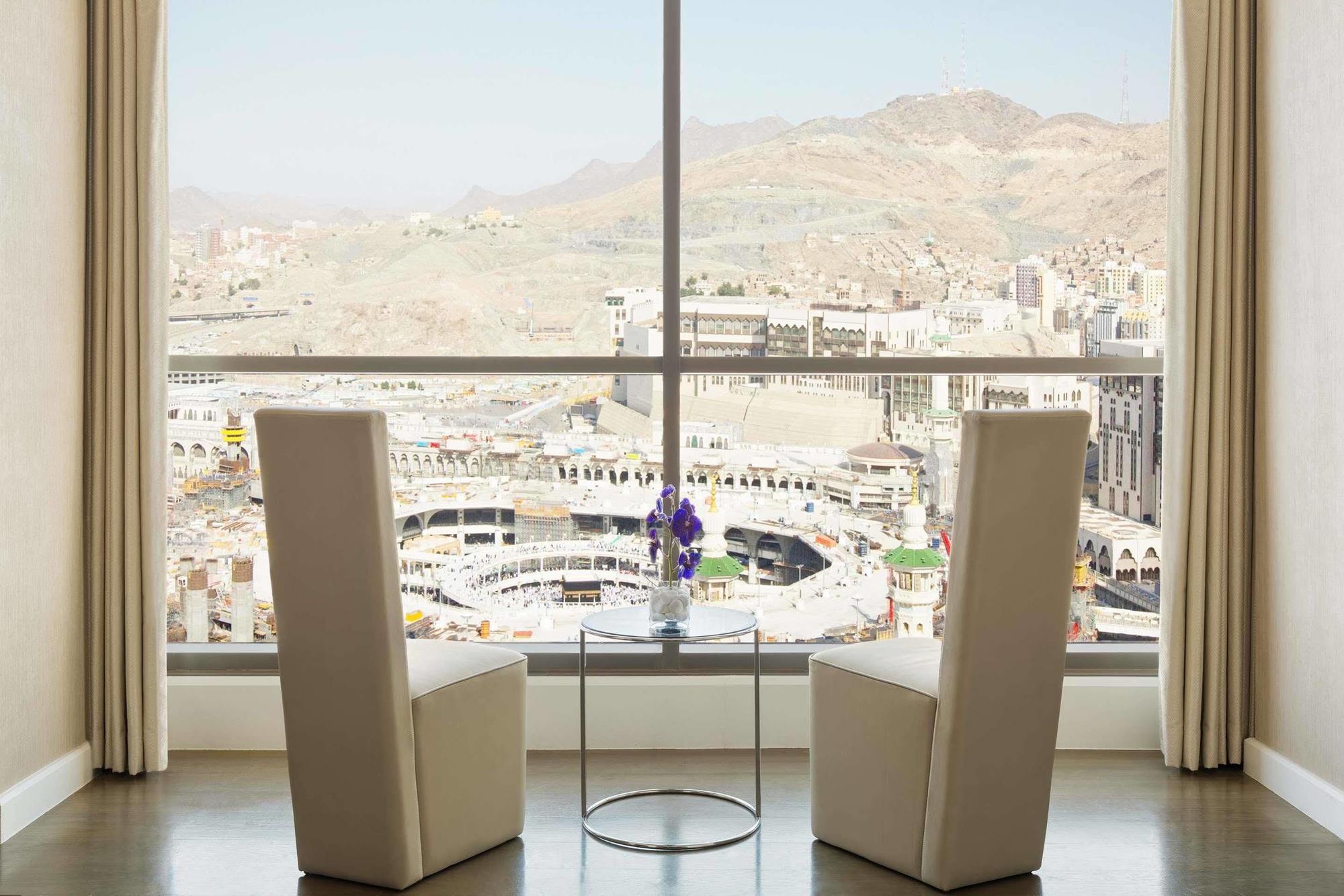 ホテル ジャバール オマール ハイアット リージェンシー マカー メッカ エクステリア 写真 The photo shows a modern indoor setting with two tall, white chairs positioned facing a large window. Through the window, there is a panoramic view of a cityscape that includes mountainous terrain and buildings, suggesting it may be a notable locatio