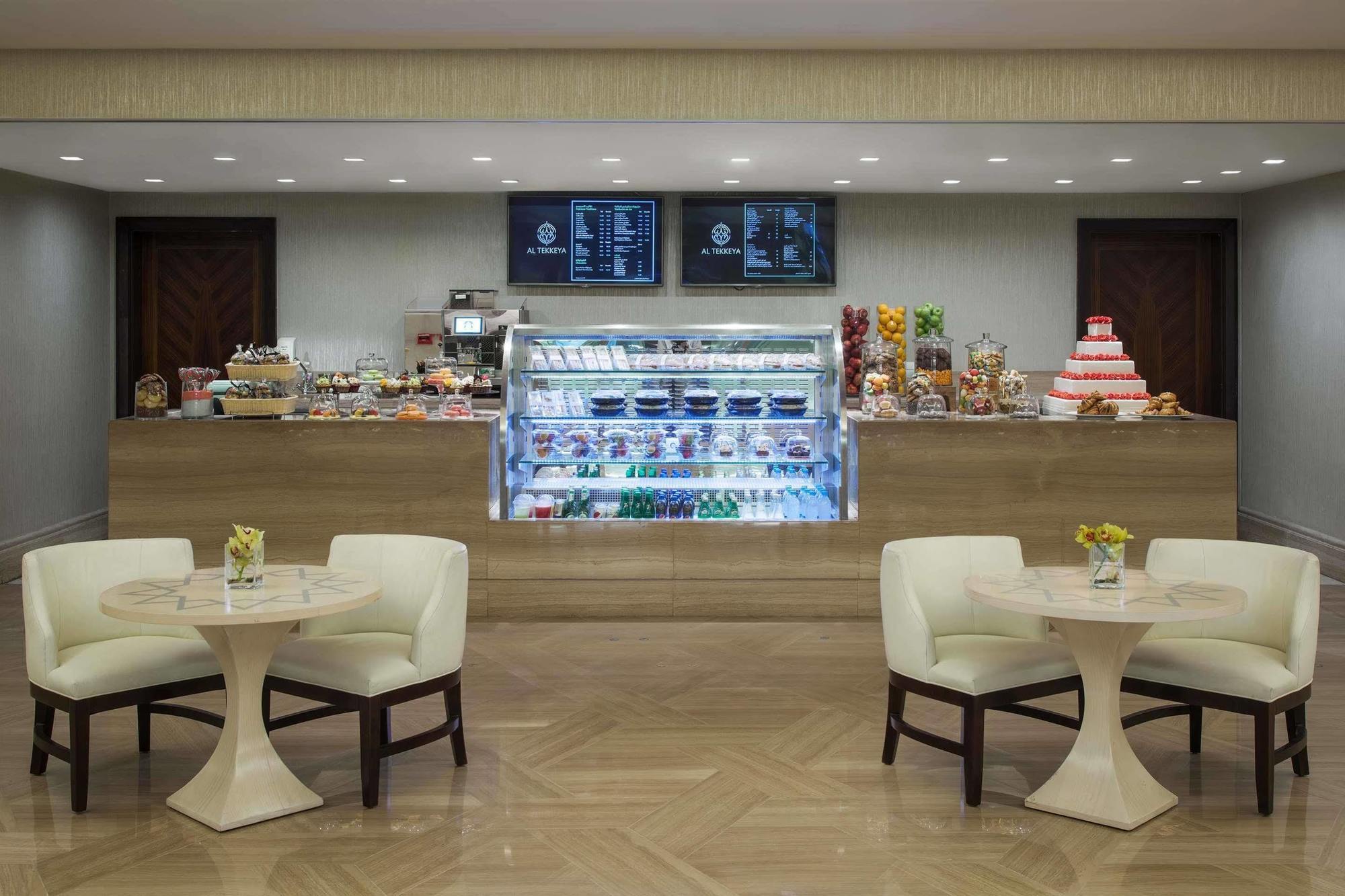 ホテル ジャバール オマール ハイアット リージェンシー マカー メッカ エクステリア 写真 The photo shows a quaint café or dessert shop interior. In the background, there's a neatly arranged counter displaying a variety of pastries and desserts, including cakes and sweets. Above the counter, two menus or screens display the offerings. In 