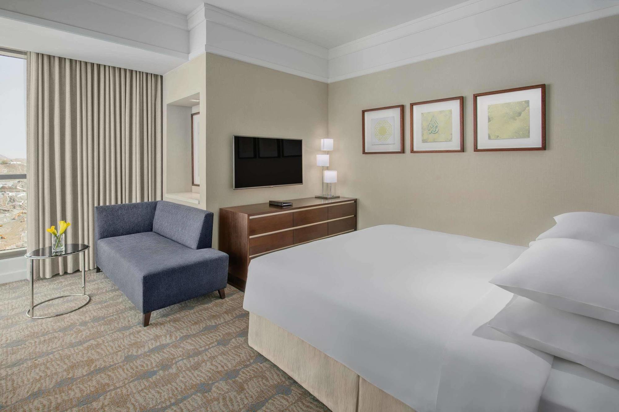 ホテル ジャバール オマール ハイアット リージェンシー マカー メッカ エクステリア 写真 The photo shows a modern hotel room featuring a neatly made bed with white linens, a small blue couch to the side, and a wooden dresser with a television mounted on the wall above it. There are two framed pieces of art on the wall near the bed, and t