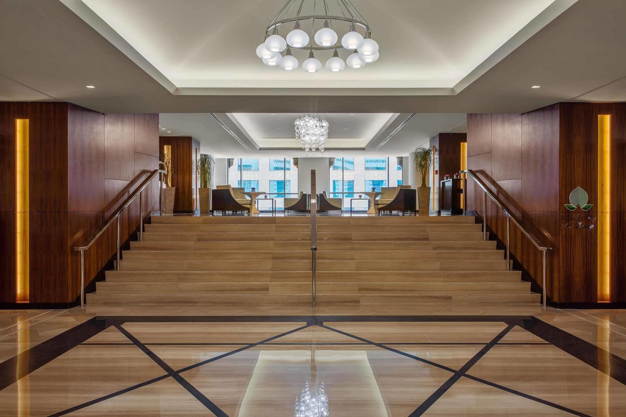 ホテル ジャバール オマール ハイアット リージェンシー マカー メッカ エクステリア 写真 The photo depicts a modern and elegant lobby area. There is a wide staircase made of wood that leads up to another level, flanked by stylish seating on both sides. The lobby features a combination of wooden and marble flooring, with a geometric patte