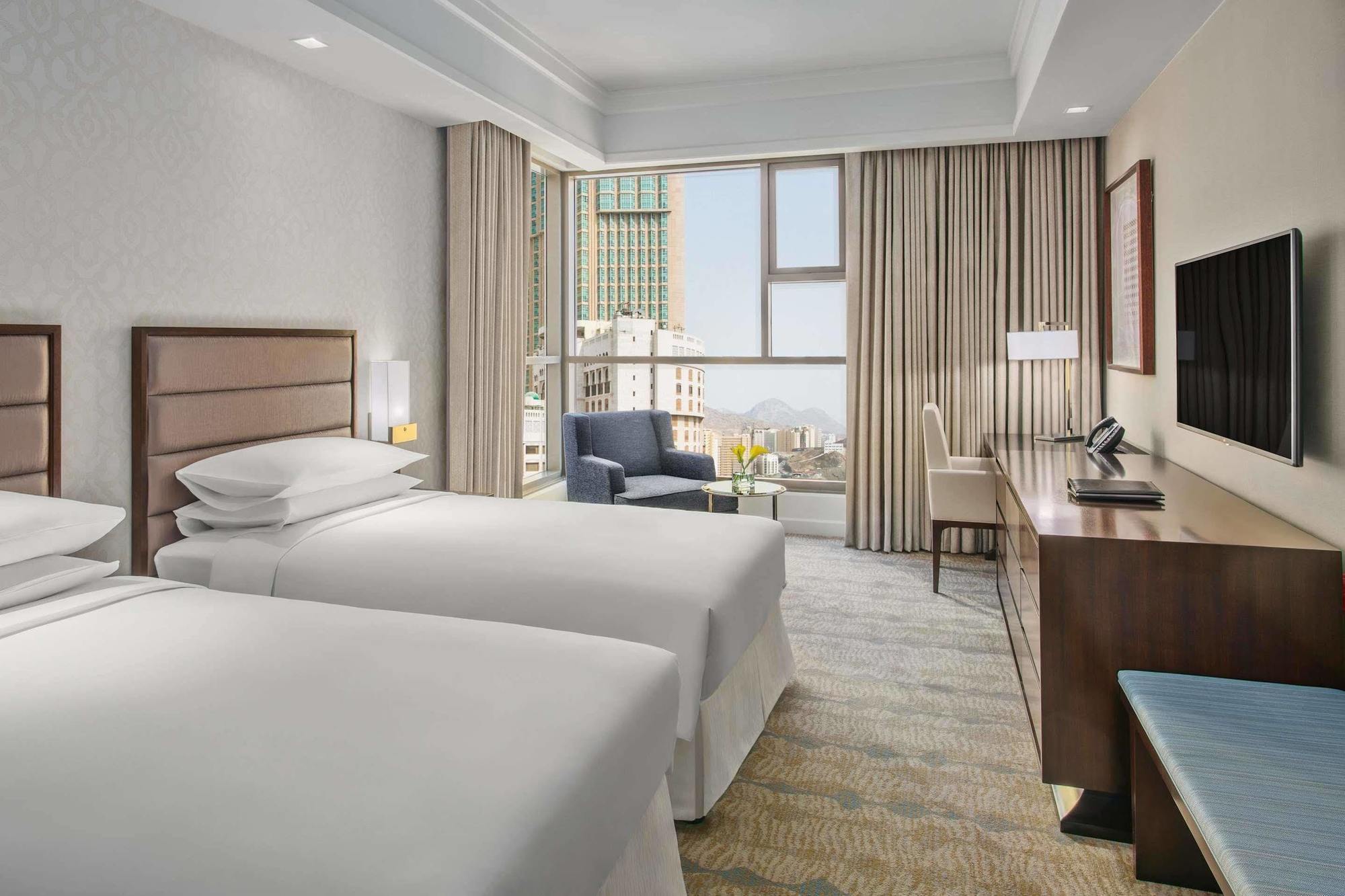 ホテル ジャバール オマール ハイアット リージェンシー マカー メッカ エクステリア 写真 The photo shows a modern hotel room featuring two single beds with white bedding. There's a spacious window allowing natural light to fill the room, which is adorned with light-colored curtains. In one corner, there's a comfortable armchair next to a
